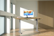 Elektrisch höhenverstellbarer Schreibtisch mit Holzoptik-Tischplatte und silbernem Metallgestell, frontal in leicht seitlicher Perspektive in einem hellen Büro mit Monitor, Tastatur und Büchern.