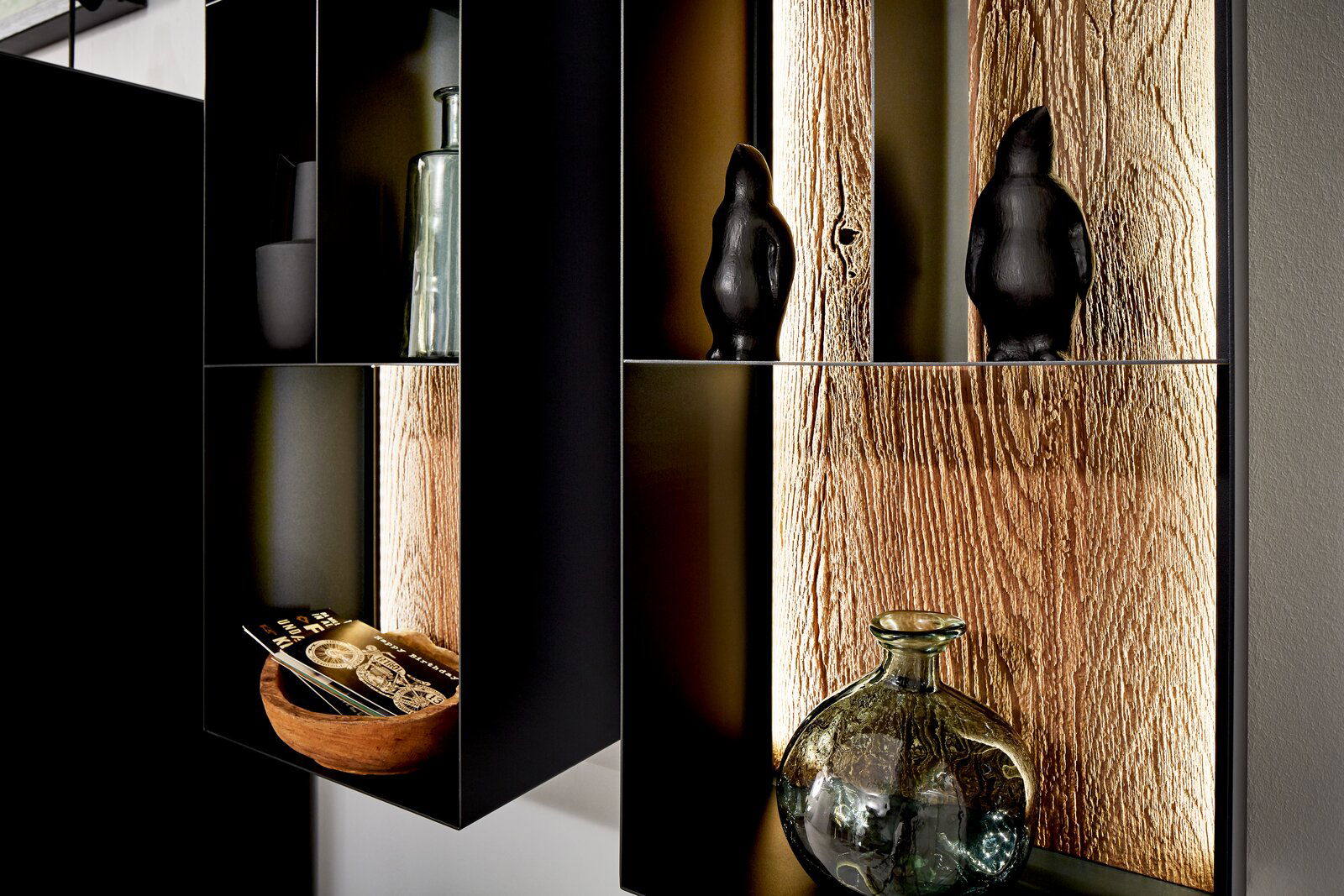 Valnatura Regal LIVINO Nahaufnahme eines modernen Regals mit dunklen Fächern, dekoriert mit Glasflaschen und schwarzen Figuren, vor einer Rückwand in Holzoptik.