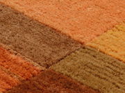 Nahaufnahme eines Teppichs mit quadratischem Muster in warmen Erdtönen wie Orange, Braun und Gelb, aus einer schrägen Perspektive fotografiert.