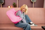 Frau sitzt auf einem Sofa mit einer großen, pinken Tasche über der Schulter, seitliche Perspektive.