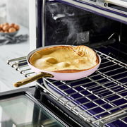 Rosa Bratpfanne mit goldfarbenem Griff im Ofen, von der Seite fotografiert, mit dampfendem Inhalt.