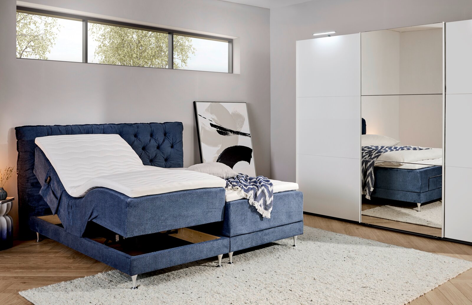 Modernes Boxspringbett in Blau mit verstellbarem Kopfteil und weißer Matratzenauflage im Schlafzimmer, seitliche Perspektive schräg von vorn