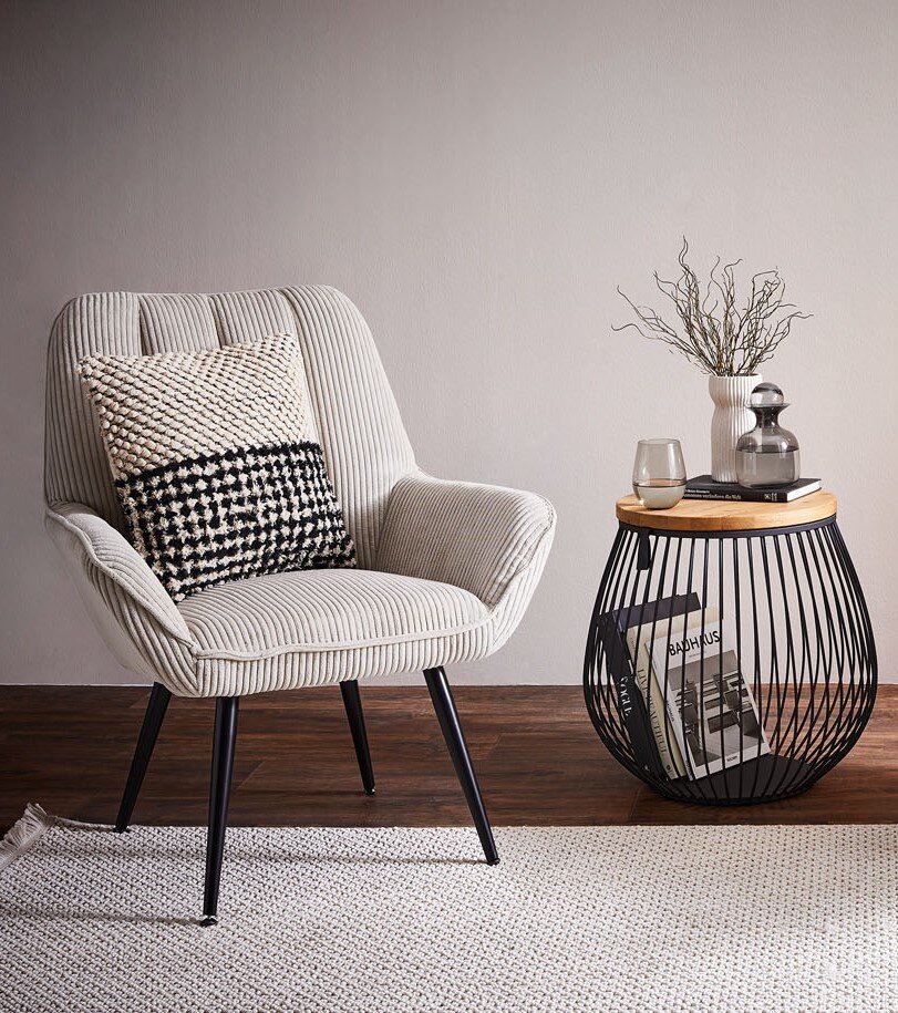 Beiger Cord-Sessel mit hoher Rückenlehne, schwarzen Metallbeinen und gemusterten Kissen, in frontaler Dreiviertelansicht neben einem runden Beistelltisch im modernen Wohnambiente