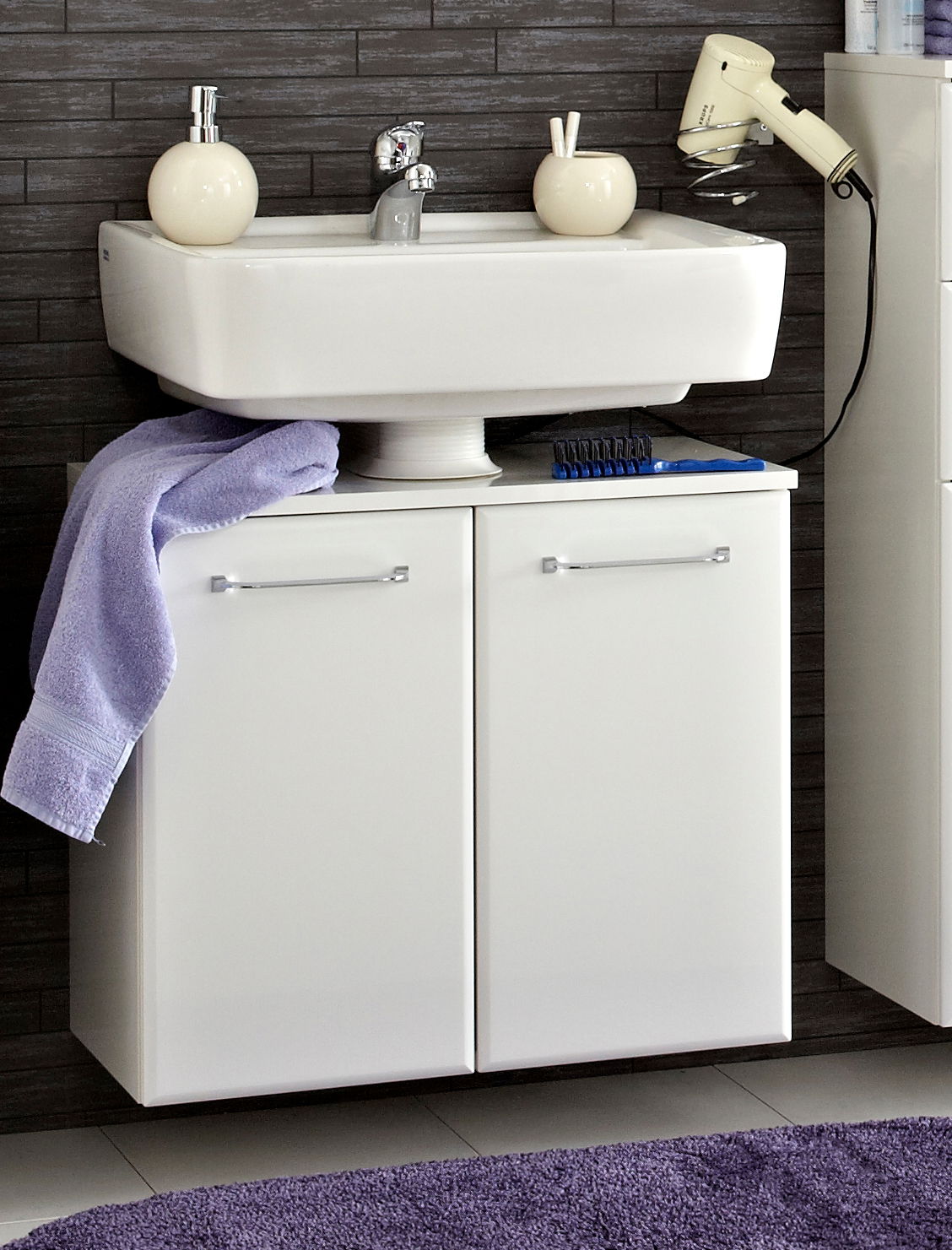 Moderner weißer Waschbeckenunterschrank mit zwei Türen, frontal fotografiert, unter einem Waschbecken montiert. Dekoriert mit einem lila Handtuch und Badezimmerutensilien.