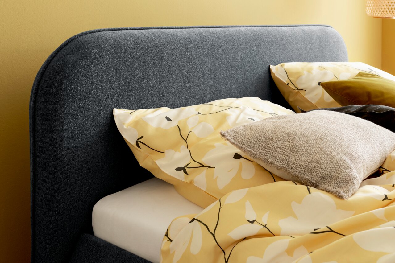 Nahaufnahme eines Polsterbetts mit grauem Kopfteil, gelber Bettwäsche mit Blumenmuster und mehreren Kissen, seitliche Perspektive