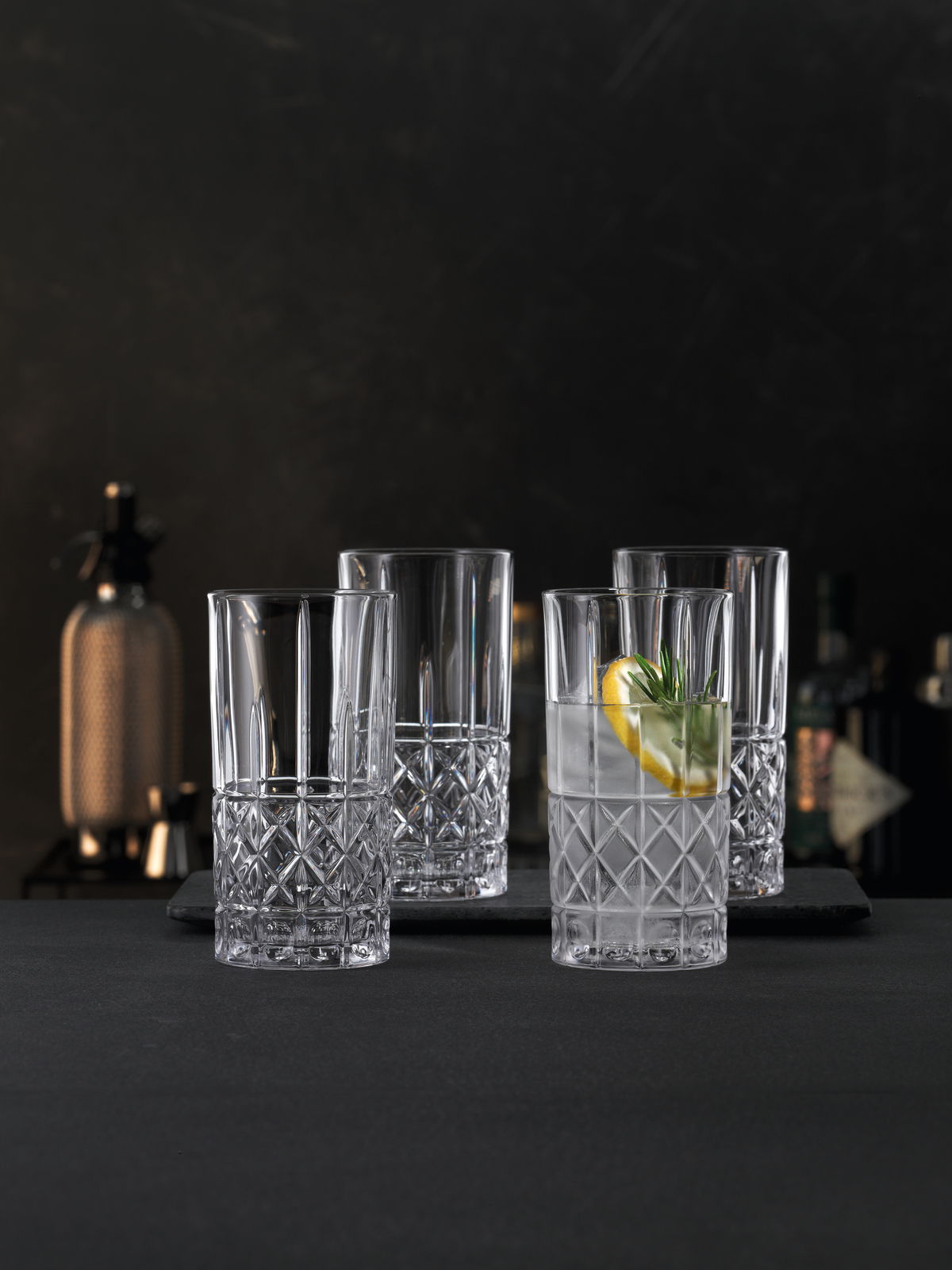 Vier elegante Trinkgläser mit Rautenmuster, gefüllt mit Getränken und Zitronenscheiben, auf einem dunklen Hintergrund. Frontale Perspektive.