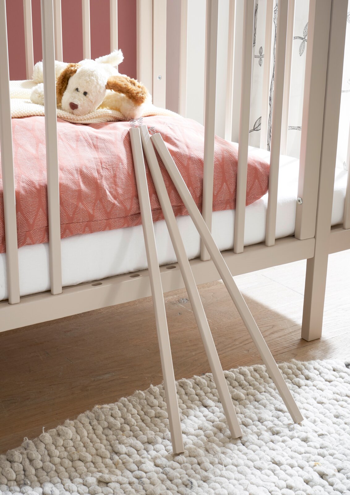 Detailaufnahme aus seitlicher Perspektive eines hellbeigen Babybetts mit rosa Bettwäsche und Kuschelhund; im Vordergrund lehnen drei abnehmbare Schlupfsprossen an den Gitterstäben, auf hellem Teppich und Holzboden.