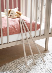 Detailaufnahme aus seitlicher Perspektive eines hellbeigen Babybetts mit rosa Bettwäsche und Kuschelhund; im Vordergrund lehnen drei abnehmbare Schlupfsprossen an den Gitterstäben, auf hellem Teppich und Holzboden.