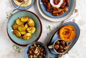 Draufsicht auf ein herbstliches Food-Arrangement mit blauen Keramikschalen: gewürzte Kartoffelhälften, gebratene Pilze, Kürbisspalten, Datteln mit cremigem Dip und geröstete Kastanien auf hellem Untergrund.