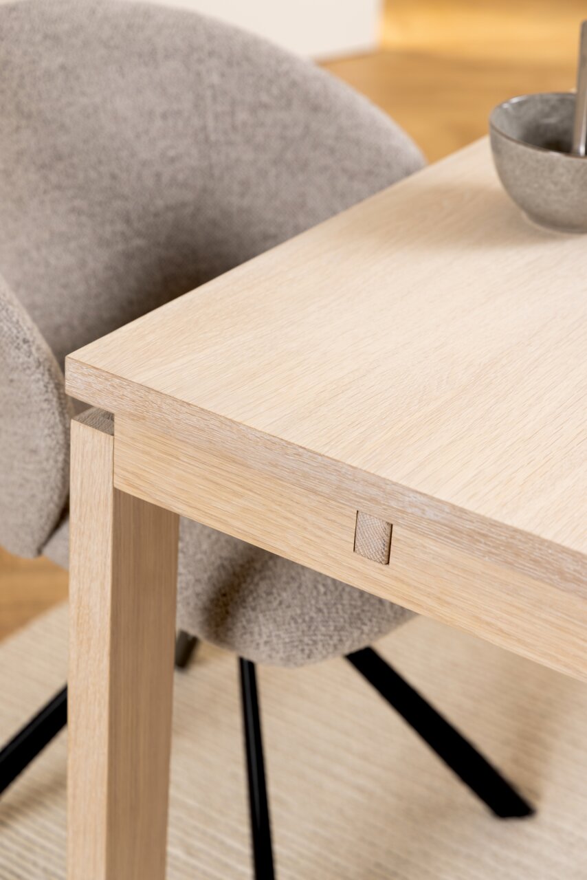 Nahaufnahme eines modernen Esstisches aus hellem Holz mit einem grauen Stuhl im Hintergrund, seitliche Perspektive.