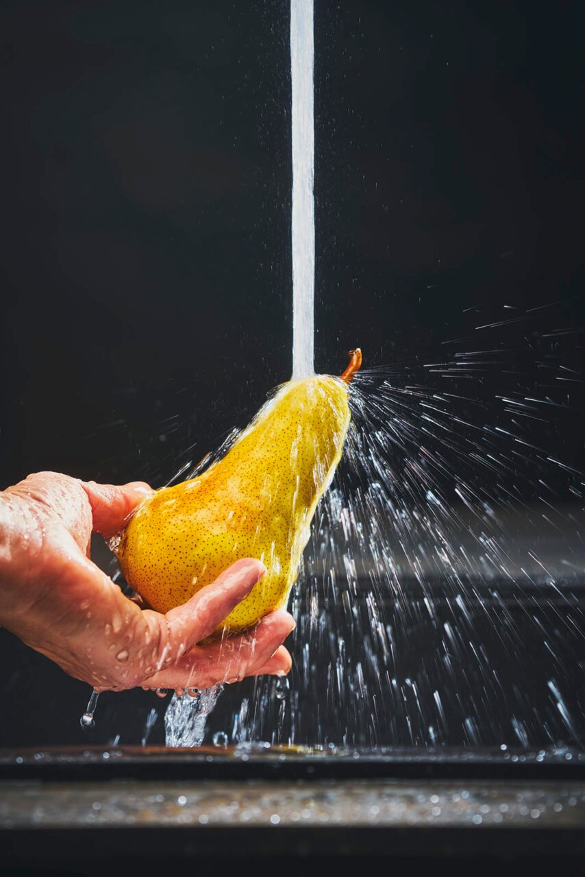 Close-up van een hand die een peer onder stromend water houdt, met opspattend water op de achtergrond.