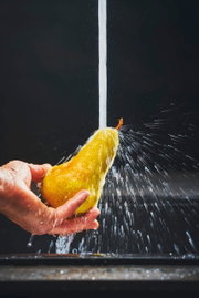 Close-up van een hand die een peer onder stromend water houdt, met opspattend water op de achtergrond.