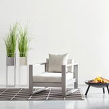 Moderner Lounge-Sessel in hellem Design, frontal fotografiert, neben zwei hohen Pflanzen und einer Feuerschale.