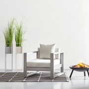Moderner Lounge-Sessel in hellem Design, frontal fotografiert, neben zwei hohen Pflanzen und einer Feuerschale.