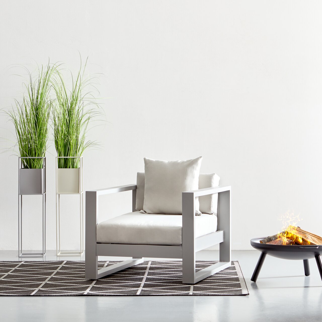 Moderne loungestoel in een lichte kleurstelling, van voren gefotografeerd, naast twee hoge planten en een vuurschaal.