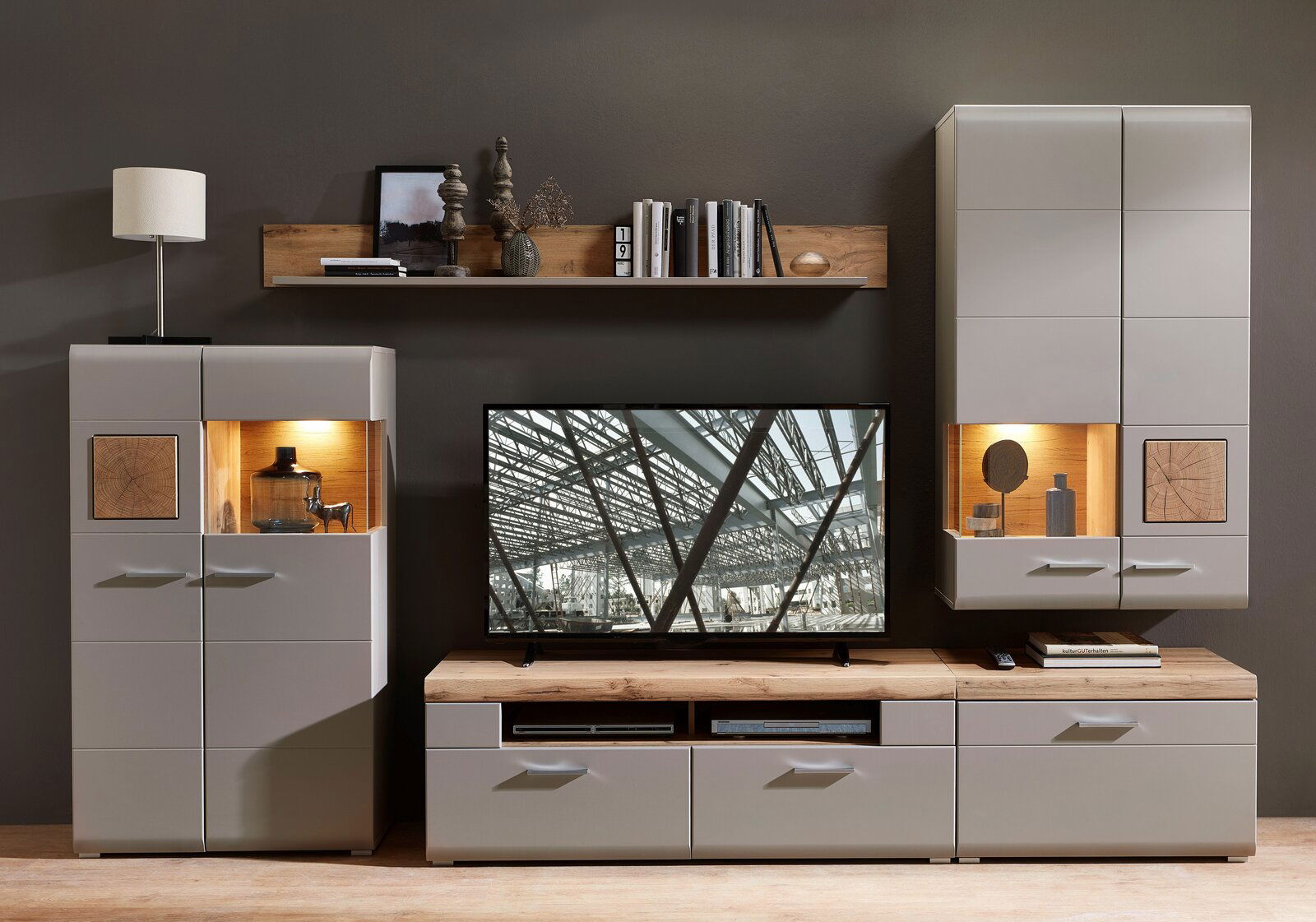 Moderne Wohnwand in grauem Design mit Holzdetails, bestehend aus einem TV-Schrank, zwei Vitrinen und einem Wandregal, frontal betrachtet.