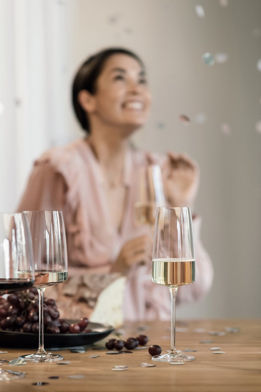 SCHOTT ZWIESEL Sekt- und Schaumweinglas VIVID SENSES Schaumweinglas auf einem Tisch mit Trauben und Käse, im Hintergrund eine feiernde Person. Perspektive: seitlich.
