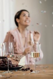 Schaumweinglas auf einem Tisch mit Trauben und Käse, im Hintergrund eine feiernde Person. Perspektive: seitlich.