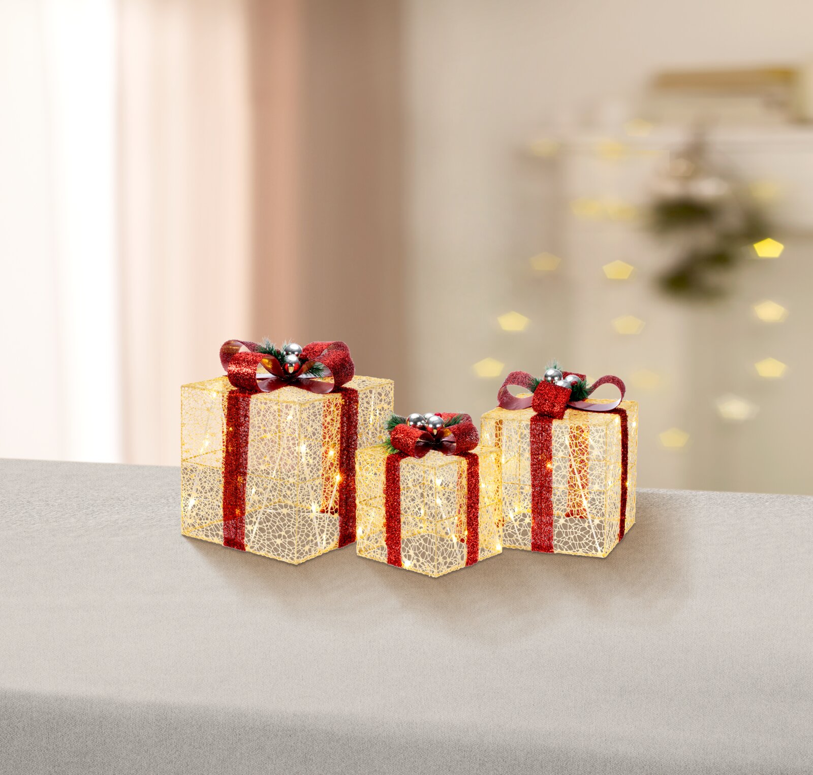 Drei dekorative LED-Geschenkboxen in Gold mit roten Glitzerbändern und Schleifen, warmweiß leuchtend, auf grauer Fläche vor unscharfem Hintergrund – Perspektive: frontale Ansicht auf Augenhöhe, leicht von oben.