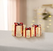 Drei dekorative LED-Geschenkboxen in Gold mit roten Glitzerbändern und Schleifen, warmweiß leuchtend, auf grauer Fläche vor unscharfem Hintergrund – Perspektive: frontale Ansicht auf Augenhöhe, leicht von oben.