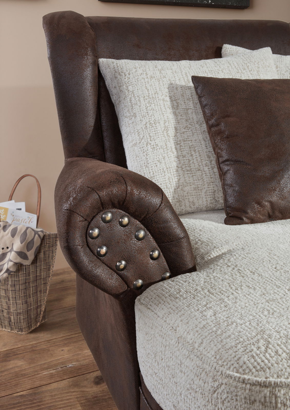Nahaufnahme eines Megaohrensessels mit brauner Lederoptik und Nieten an der Armlehne, kombiniert mit hellen Kissen. Perspektive von der Seite.
