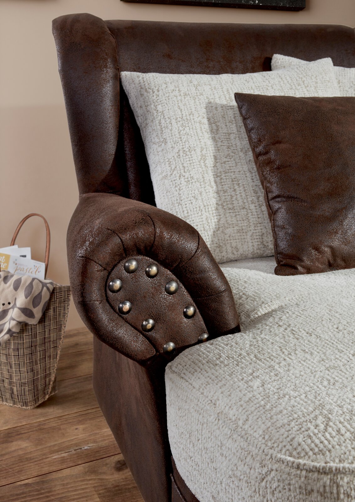 Nahaufnahme eines Megaohrensessels mit brauner Lederoptik und Nieten an der Armlehne, kombiniert mit hellen Kissen. Perspektive von der Seite.