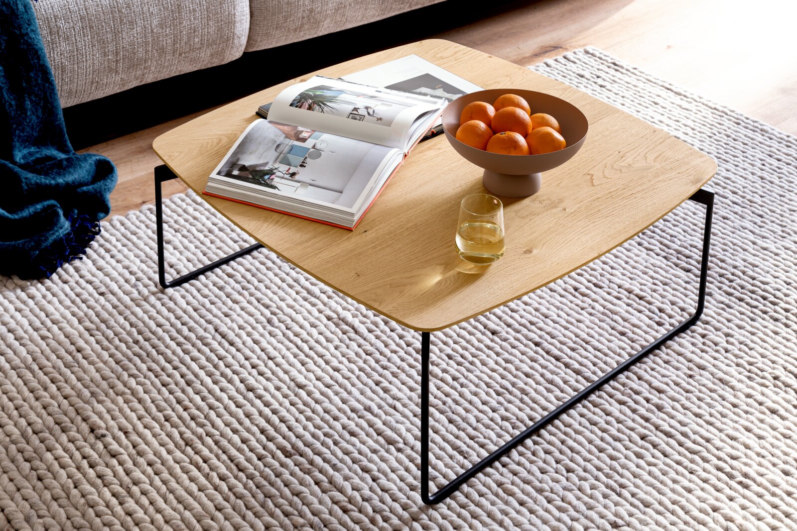 Vierkante salontafel met licht eikenhouten blad en zwart metalen frame, gestyled met open fotoboek, schaal met sinaasappels en glas op een grof geweven vloerkleed, gezien schuin van boven.