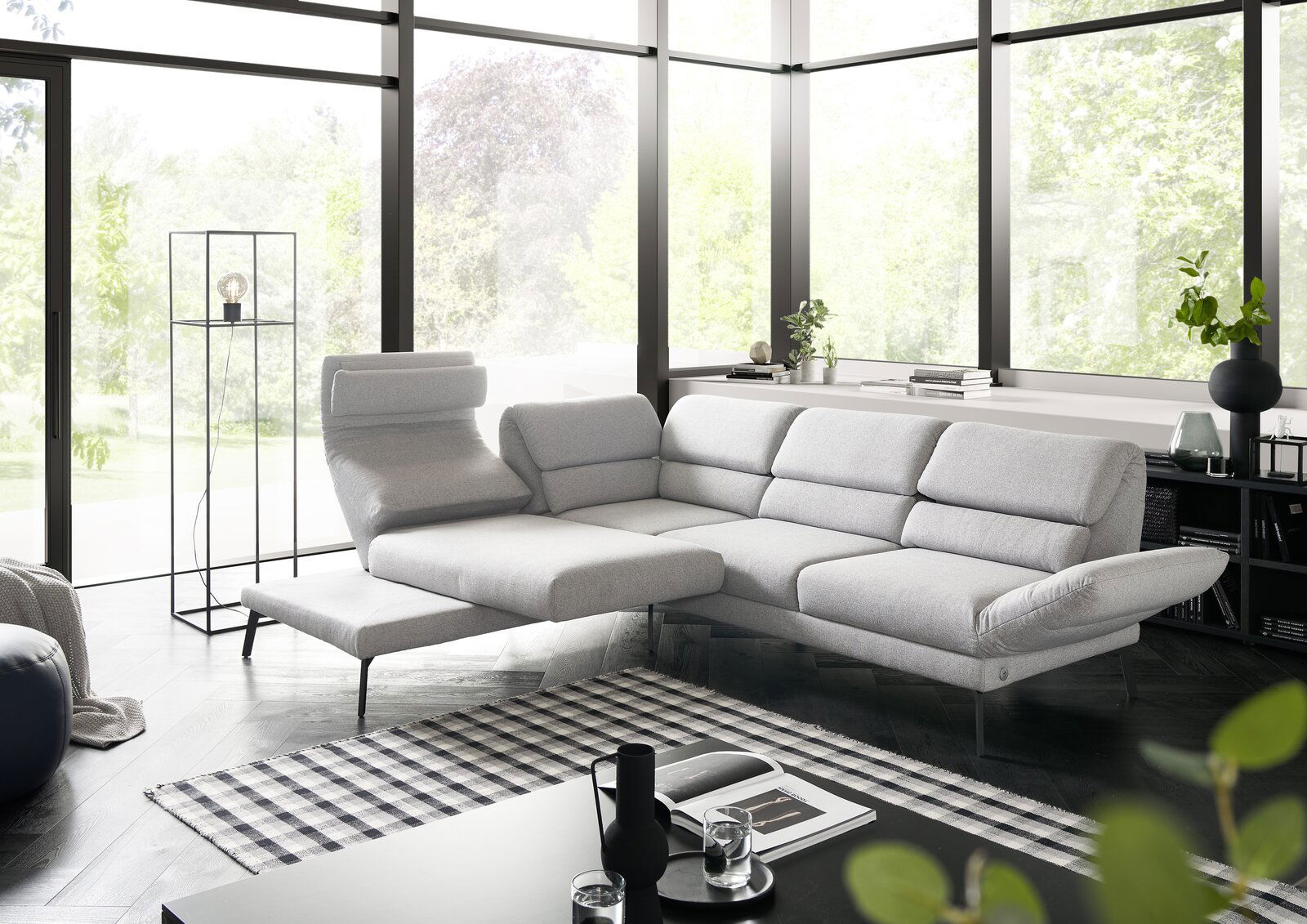 Moderne graue Eckgarnitur in einem hellen Wohnzimmer mit großen Fenstern, seitliche Perspektive.