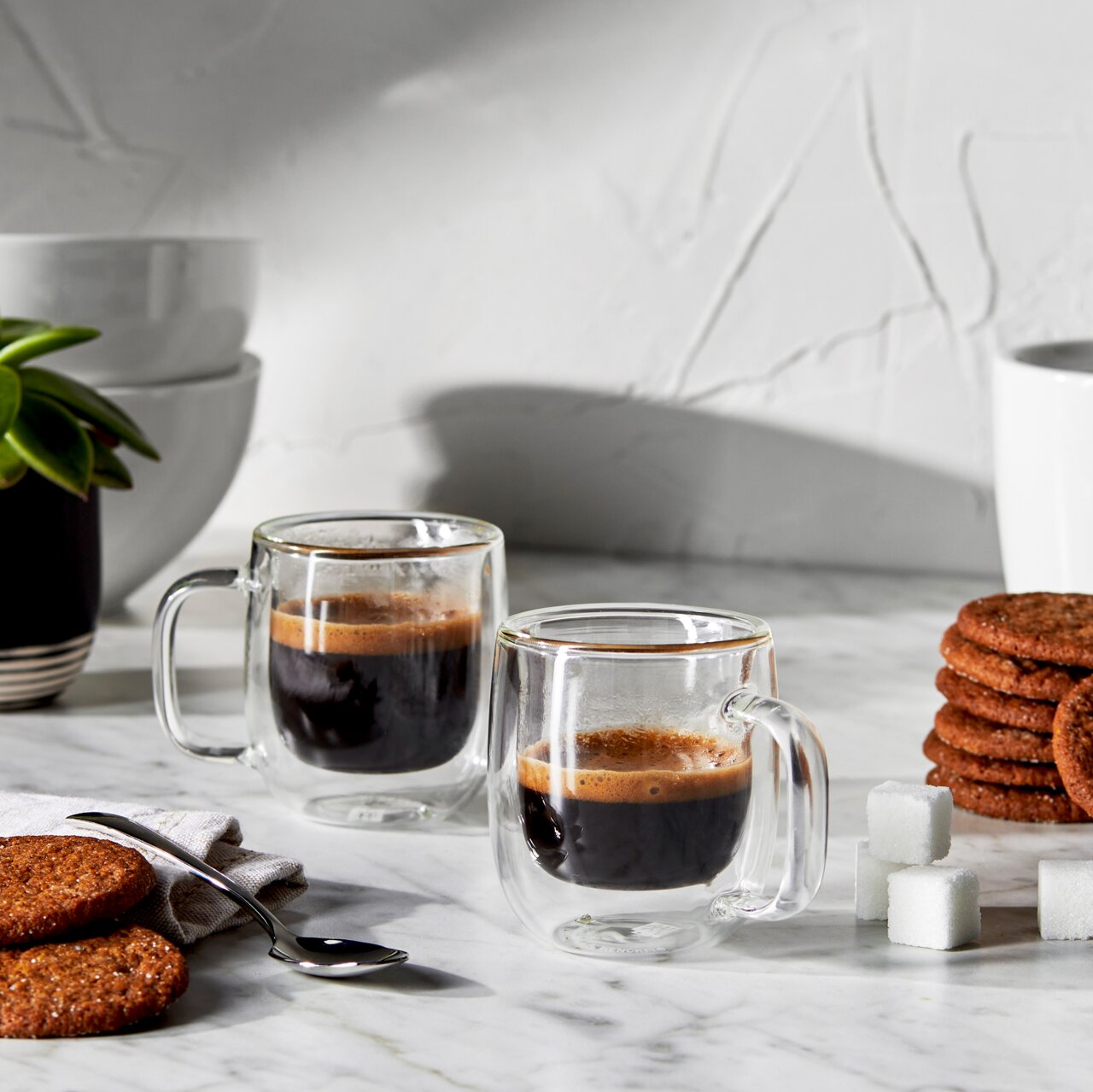 Twee dubbelwandige espressokopjes met zwarte koffie op een marmeren tafel, omringd door koekjes, suikerklontjes en een plant, zijaanzicht.