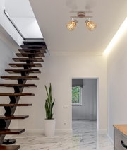 Moderne Deckenleuchte mit zwei schwenkbaren Spots und goldfarbenen Drahtkäfig-Schirmen, warm leuchtend, in einem hellen Flur neben einer Holztreppe – Ansicht von unten nach oben zur Decke.