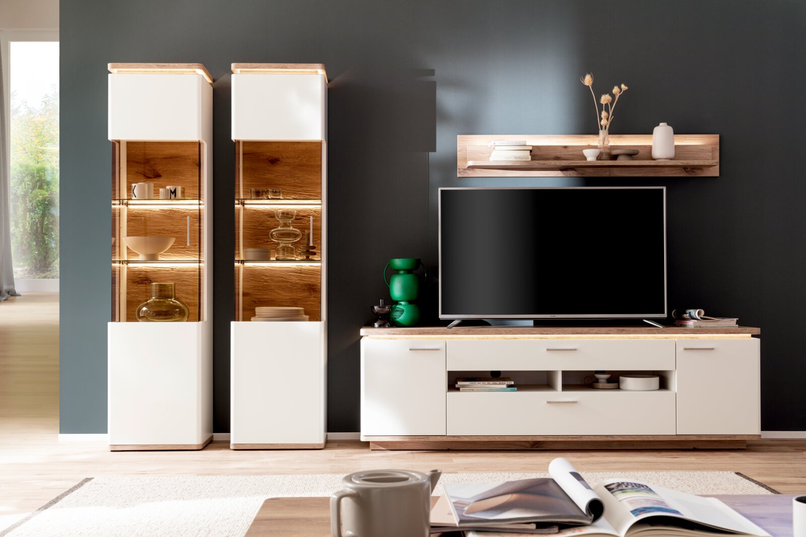 Wohnzimmer-Wohnwand in Weiß und Holzoptik mit zwei beleuchteten Vitrinen, TV-Lowboard und Wandboard vor dunkelgrauer Wand, frontal aus Augenhöhe fotografiert
