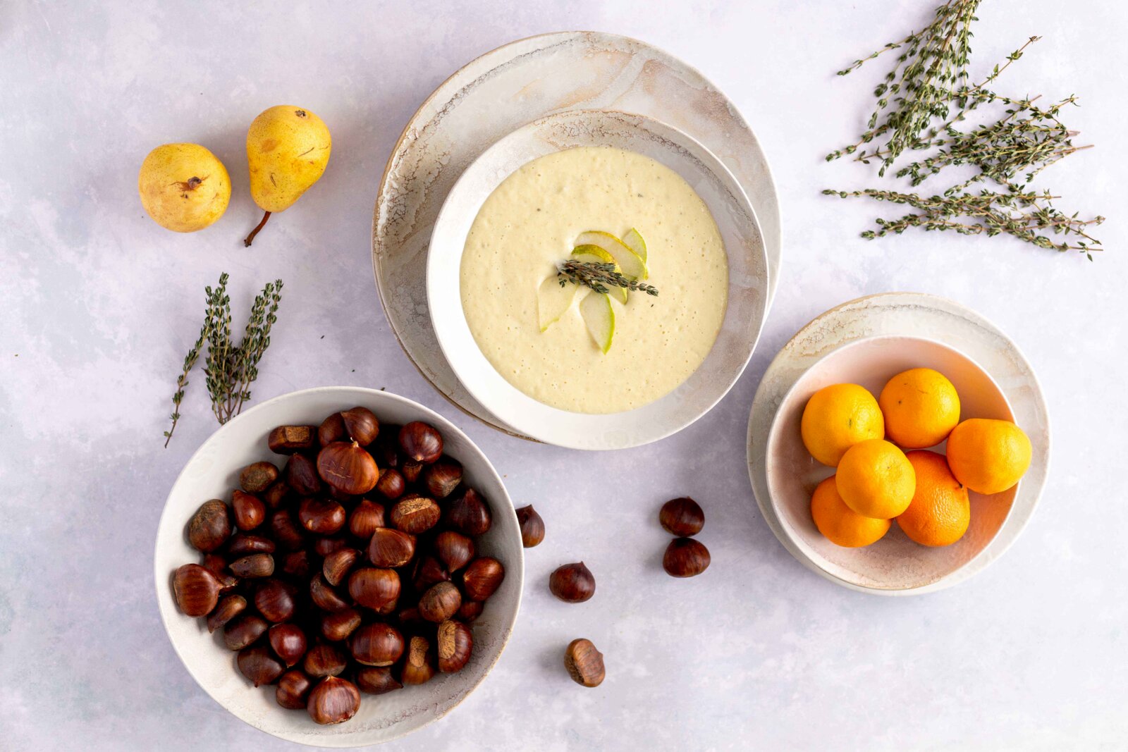 Mäser Tellerset 18-tlg. OSSIA Draufsicht auf eine Schale mit cremiger Suppe, garniert mit Birnenscheiben und Thymian, umgeben von Birnen, Kastanien und Orangen auf hellem Hintergrund