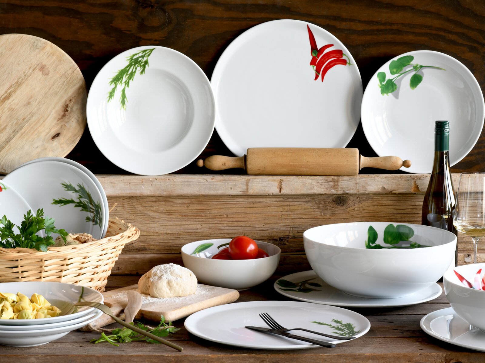Rustikales Stillleben mit weißem Porzellan-Geschirrset (Teller, Schalen, große Servierschüssel) mit Kräuter- und Chili-Motiven auf Holztisch, dazu Nudelholz, Tomaten, Teig, Kräuter und Weinflasche; Perspektive: frontal mit leicht erhöhter Ansicht.