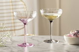Zwei elegante Champagnerschalen auf einem Marmortisch, eine in Rosa und eine in Grau, mit einer seitlichen Perspektive.