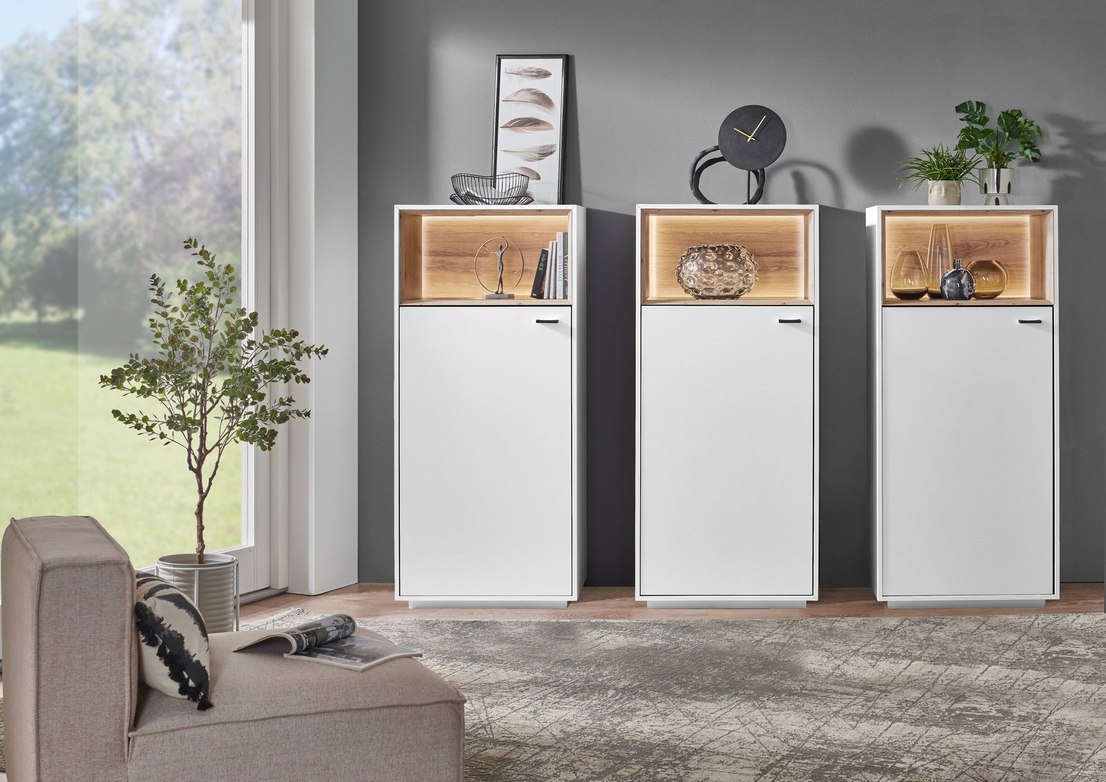 Drei moderne Highboard-Vitrinen in Weiß mit Holzdetails, dekoriert mit Pflanzen und Kunstobjekten, in einem stilvollen Wohnzimmer mit Blick auf den Garten.