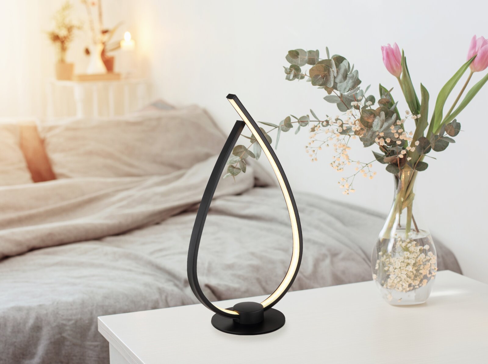 Moderne schwarze LED-Tischleuchte in geschwungener Tropfenform mit warmweißem Licht auf weißem Nachttisch neben Glasvase mit Blumen im Schlafzimmer, Perspektive: frontal in leichter Seitenansicht