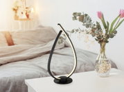 Moderne schwarze LED-Tischleuchte in geschwungener Tropfenform mit warmweißem Licht auf weißem Nachttisch neben Glasvase mit Blumen im Schlafzimmer, Perspektive: frontal in leichter Seitenansicht