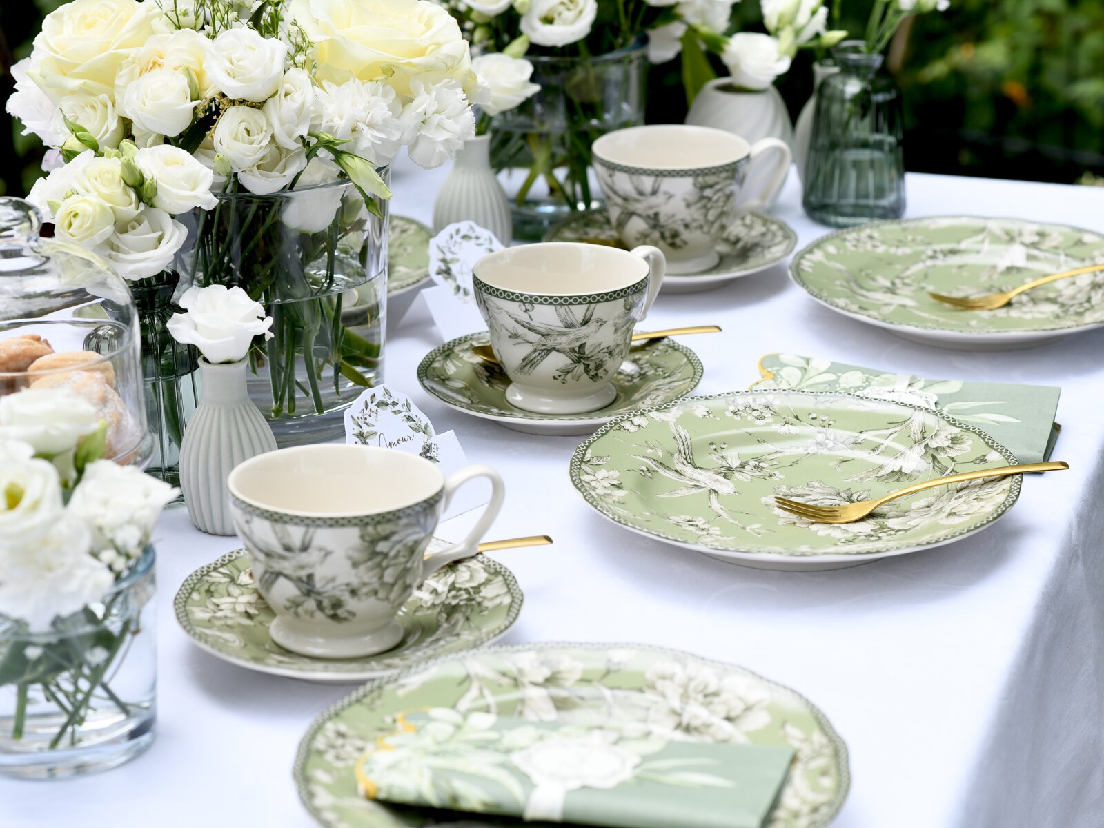 Schräg von oben fotografierte, elegant gedeckte Tafel mit grün-weiß gemustertem Porzellan (Teller und Tassen), goldfarbenem Besteck, passenden Servietten und weißen Rosen in Glasvasen.
