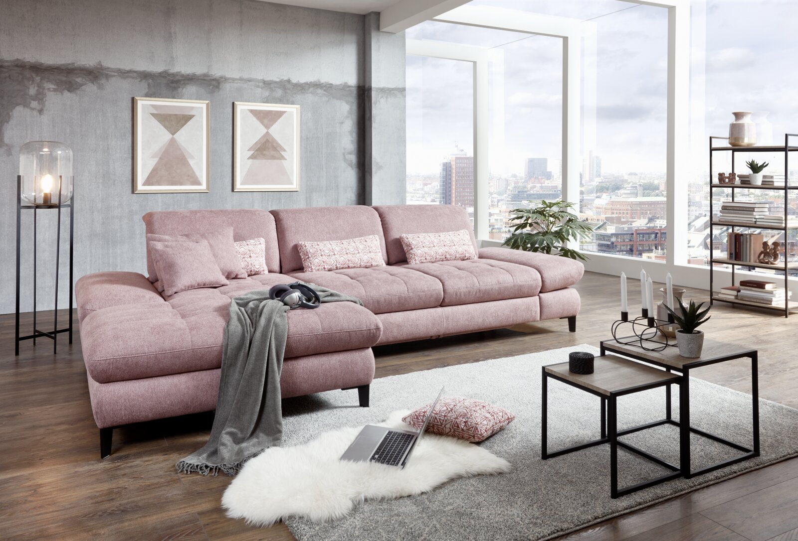 Grote, moderne hoekbank in roze met kussens, in een stijlvolle woonkamer met grote ramen en uitzicht op de stad, gefotografeerd vanuit een zijdelingse hoek.