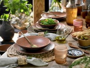 Schräg von oben fotografierter, rustikal gedeckter Esstisch mit roséfarbenem Keramikgeschirr, goldfarbenem Besteck, Salat in Schale, Olivenölkaraffe, Oliven, Brotstangen, Gewürzschale und farbigen Gläsern in warmem Tageslicht.