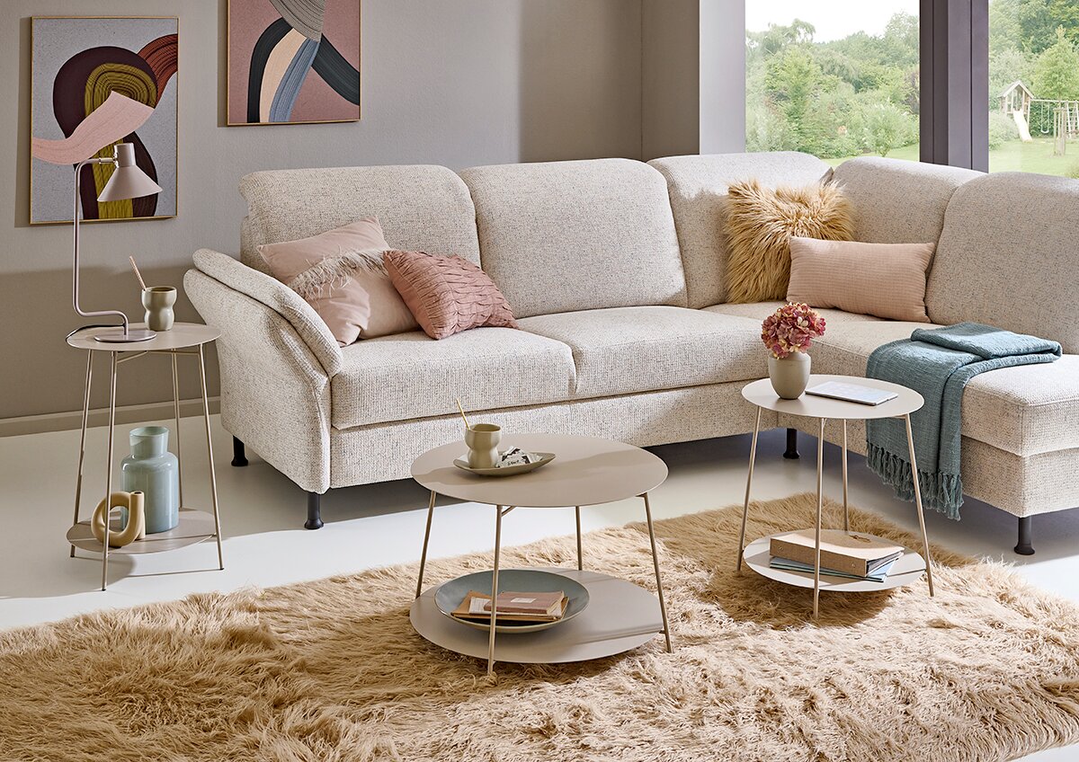 Zurbrüggen Beistelltisch Modernes Wohnzimmer mit hellbeigem, stoffbezogenem Ecksofa (L-Form) mit Kissen und Tagesdecke sowie runden Beistelltischen auf flauschigem Teppich; Perspektive: frontale, leicht schräge Ansicht von links auf das Sofa.