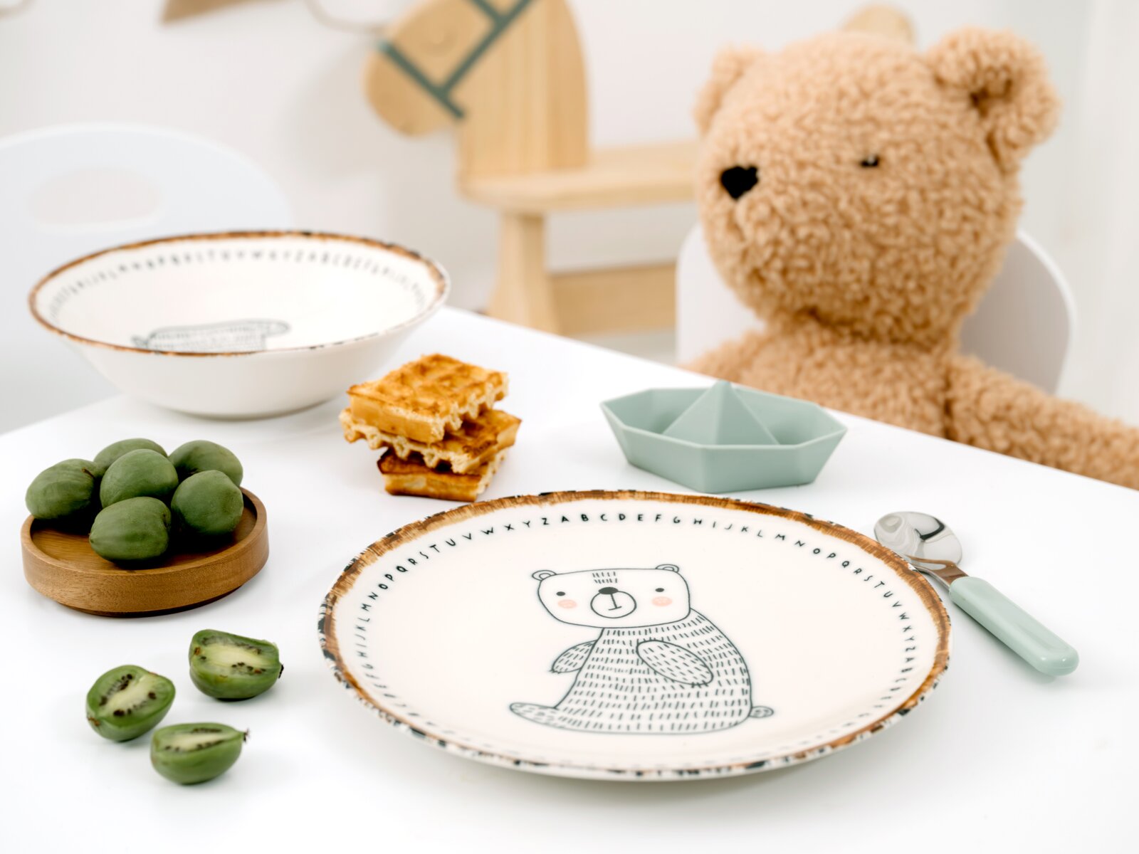 Kinder-Geschirrset mit Bärenmotiv, bestehend aus Teller, Schale, Löffel und mintgrünem Snackschälchen, schräg von oben auf einem gedeckten Kindertisch fotografiert