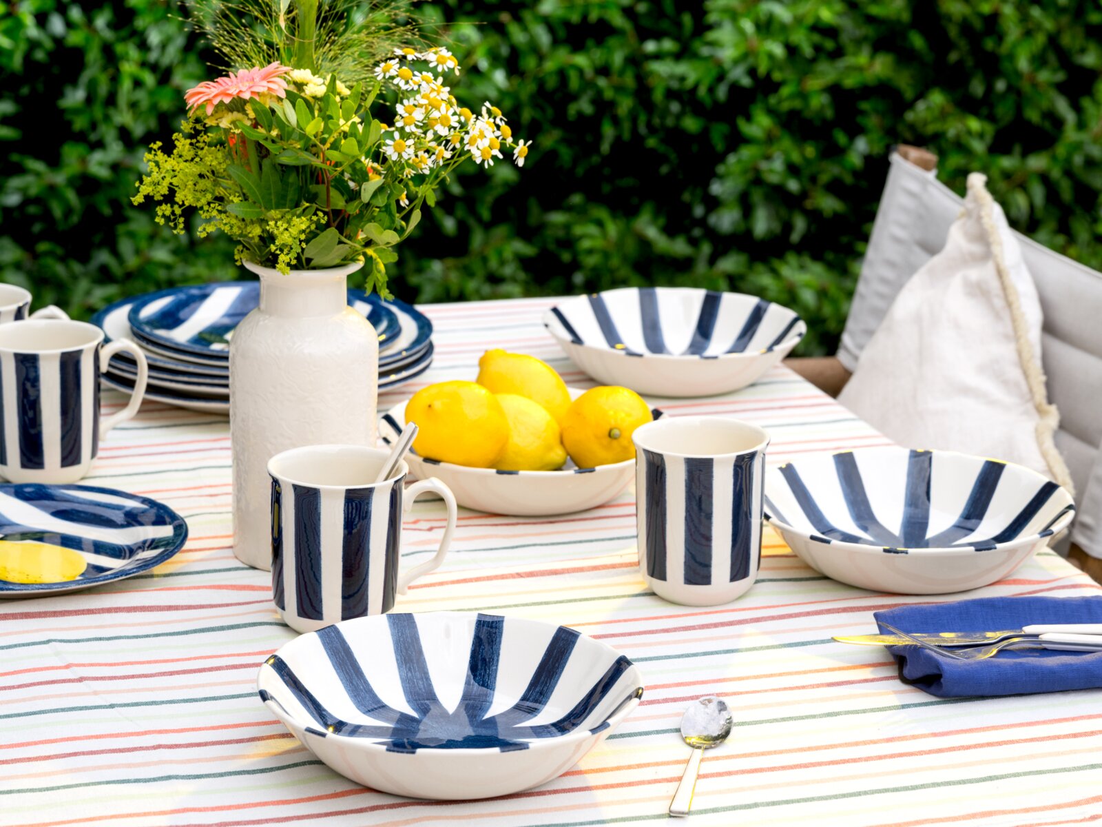 Schräg von vorne fotografierte Outdoor-Tischszene mit blau-weiß gestreiftem Geschirr (Schalen, Teller, Becher), weißer Vase mit Blumenstrauß und Schale mit Zitronen auf gestreifter Tischdecke im Garten.