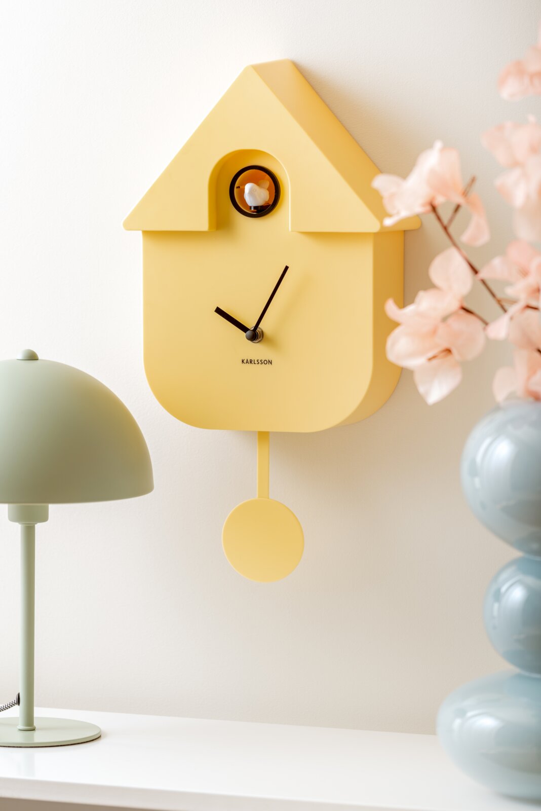 Gele wandklok in de vorm van een huisje met slinger en een vogeltje, zijaanzicht