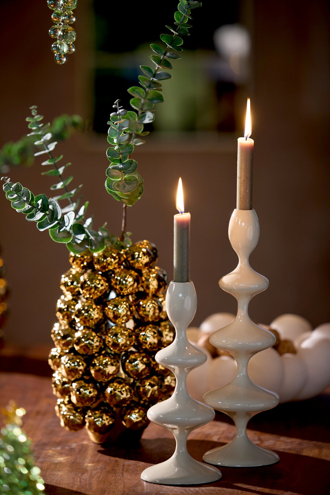 Zwei elegante, weiße Kerzenständer mit geschwungenem Design, auf einem Tisch arrangiert, mit brennenden Kerzen und dekorativen Elementen im Hintergrund. Seitliche Perspektive.