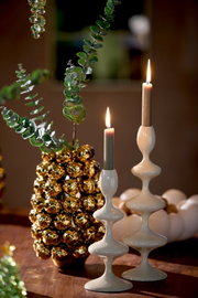 Zwei elegante, weiße Kerzenständer mit geschwungenem Design, auf einem Tisch arrangiert, mit brennenden Kerzen und dekorativen Elementen im Hintergrund. Seitliche Perspektive.
