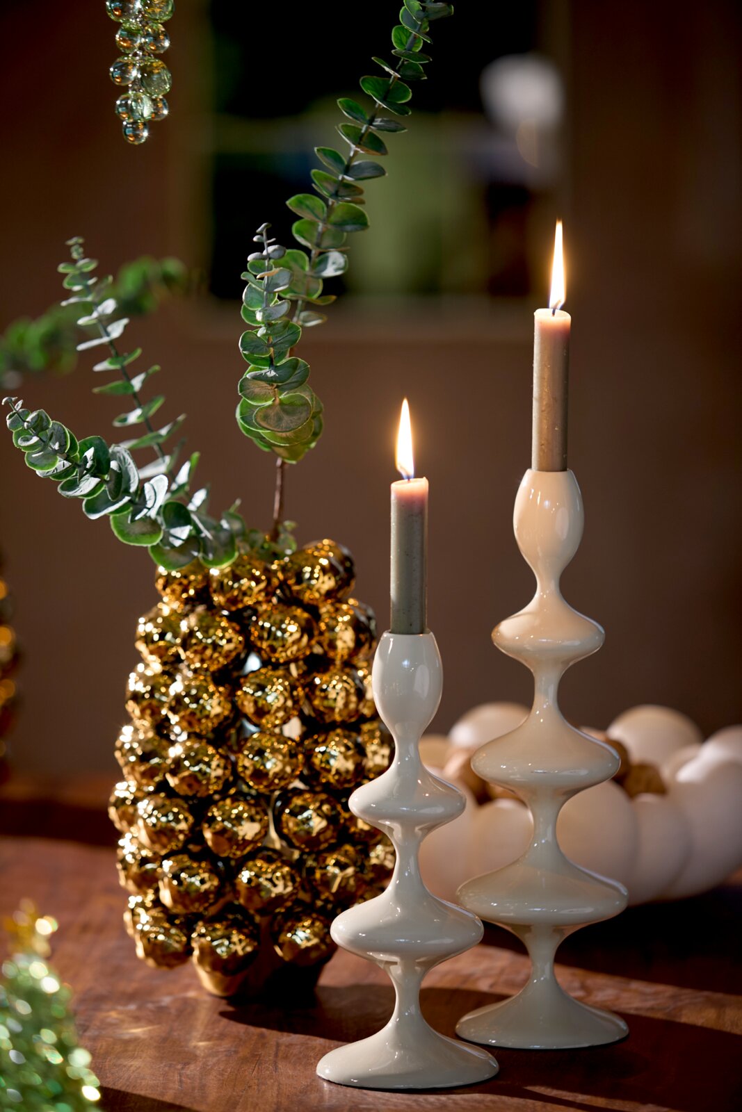 Zwei elegante, weiße Kerzenständer mit geschwungenem Design, auf einem Tisch arrangiert, mit brennenden Kerzen und dekorativen Elementen im Hintergrund. Seitliche Perspektive.