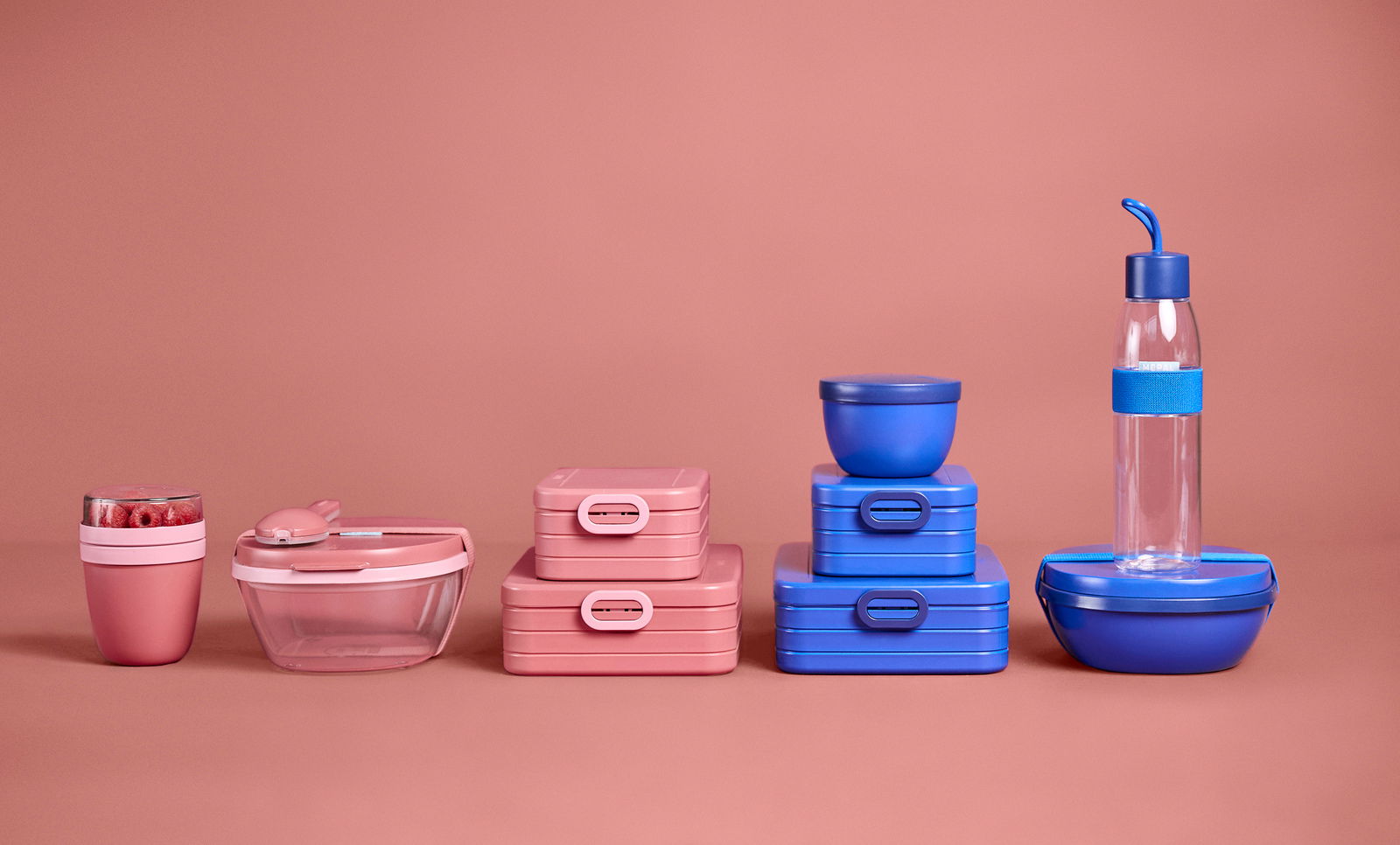 Verschiedene Lunchboxen und Behälter in Rosa und Blau, frontal auf einem rosa Hintergrund präsentiert.