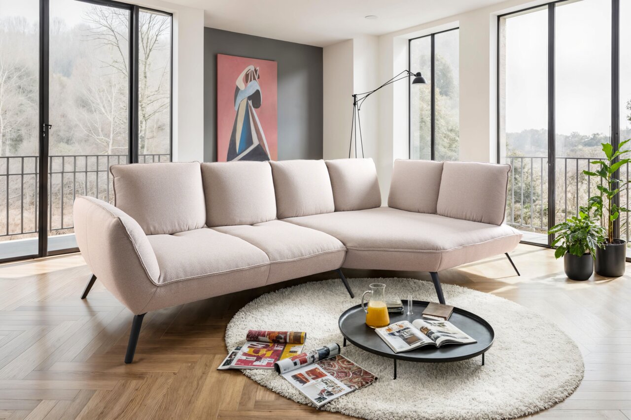 Moderne Longchairkombination in einem hellen Wohnzimmer mit großen Fenstern, die eine Aussicht auf die Natur bieten. Die Perspektive zeigt das Sofa von vorne, umgeben von einem runden Teppich und einem Couchtisch mit Zeitschriften und einem Krug Orangensa