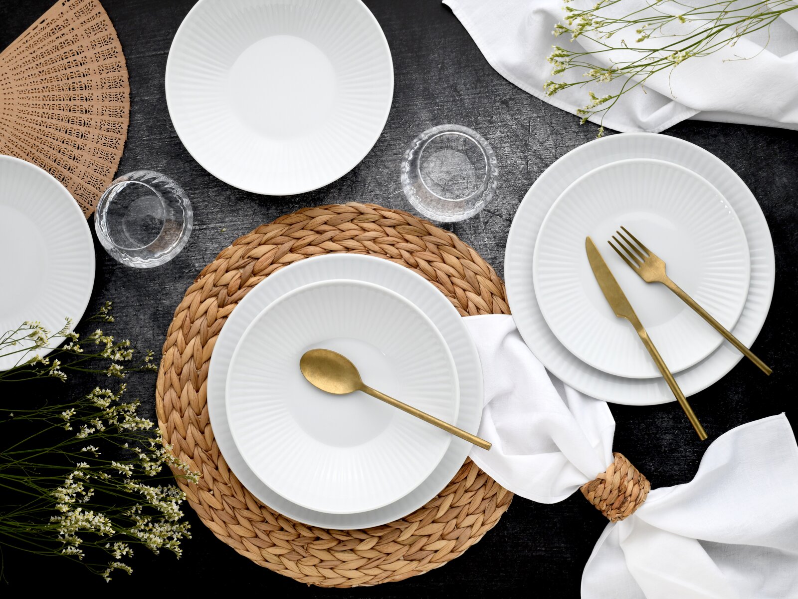 Draufsicht auf ein elegant gedecktes Tischarrangement mit weißen, geriffelten Porzellantellern und -schalen, goldfarbenem Besteck, geflochtenem Platzset, Glasuntersetzern und weißer Serviette auf dunklem Untergrund.
